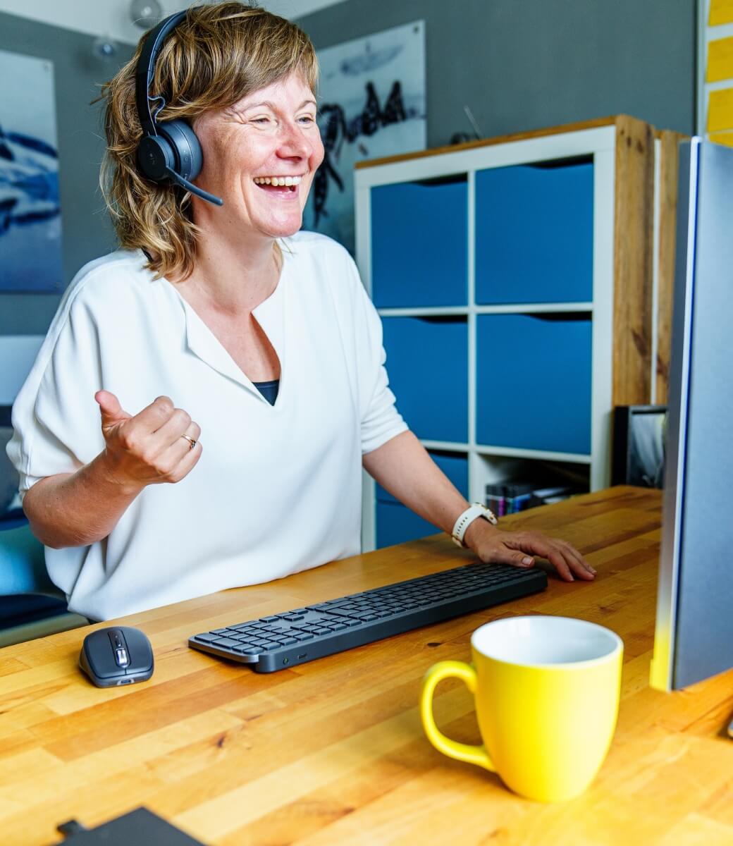 Frau mit Kopfhörer steht lächelnd an einer Tastatur am Schreibtisch auf dem eine gelbe Tasse steht